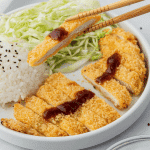 Air fryer chicken katsu on a white plate and chopsticks holding a piece.