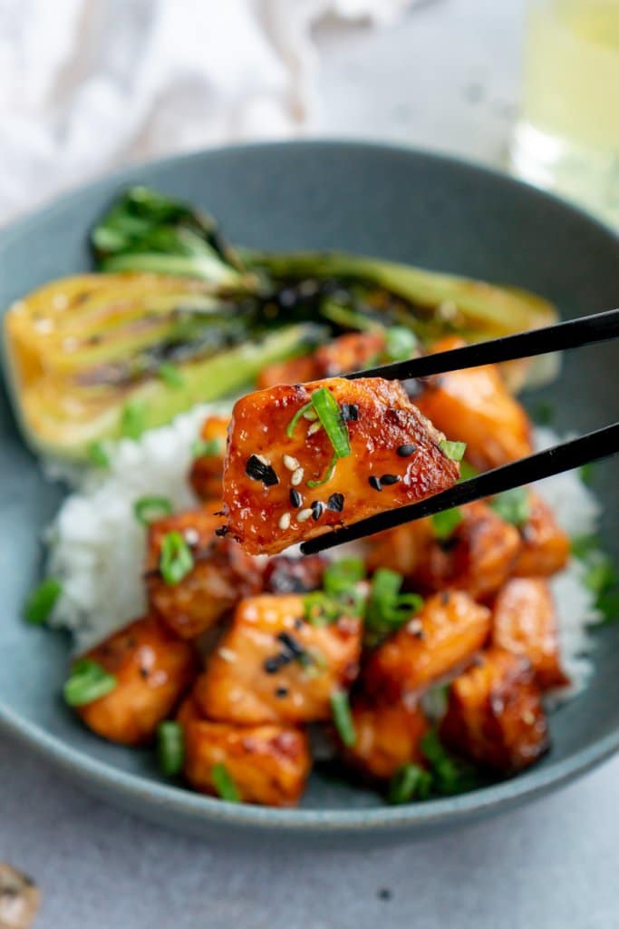 A pair of chopsticks holding one miso salmon bite in front of rice and bok choy.