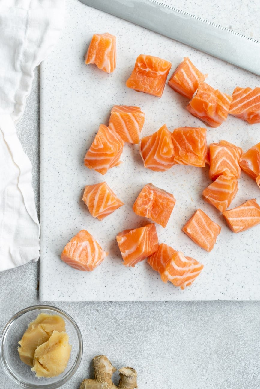 Salmon cubes on a white cutting board with a knife and fresh ginger.