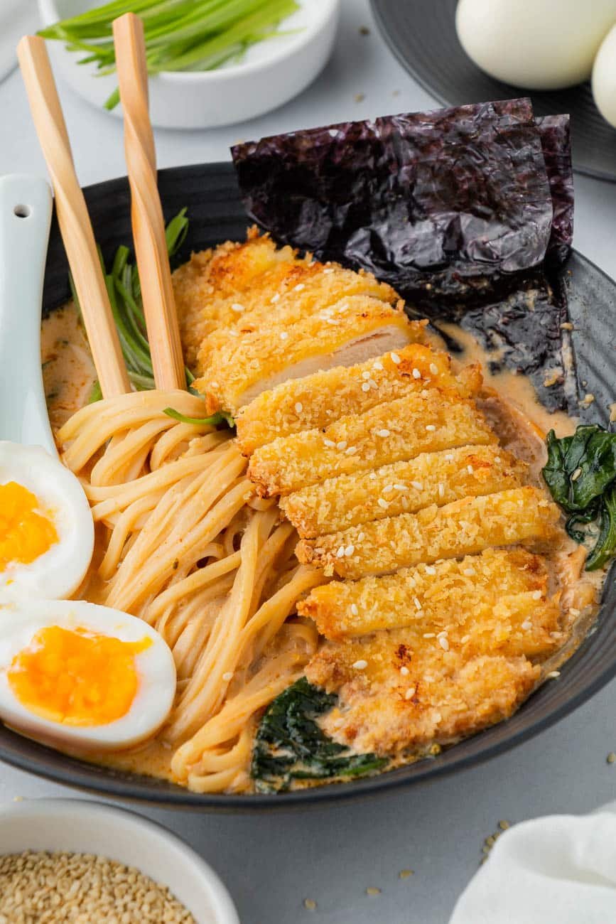 Close up of chicken katsu ramen in a black bowl with chopsticks, soft boiled eggs, and nori sheets.