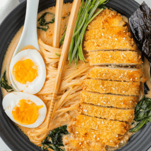 chicken katsu ramen in a black bowl with soft boiled eggs and chopsticks