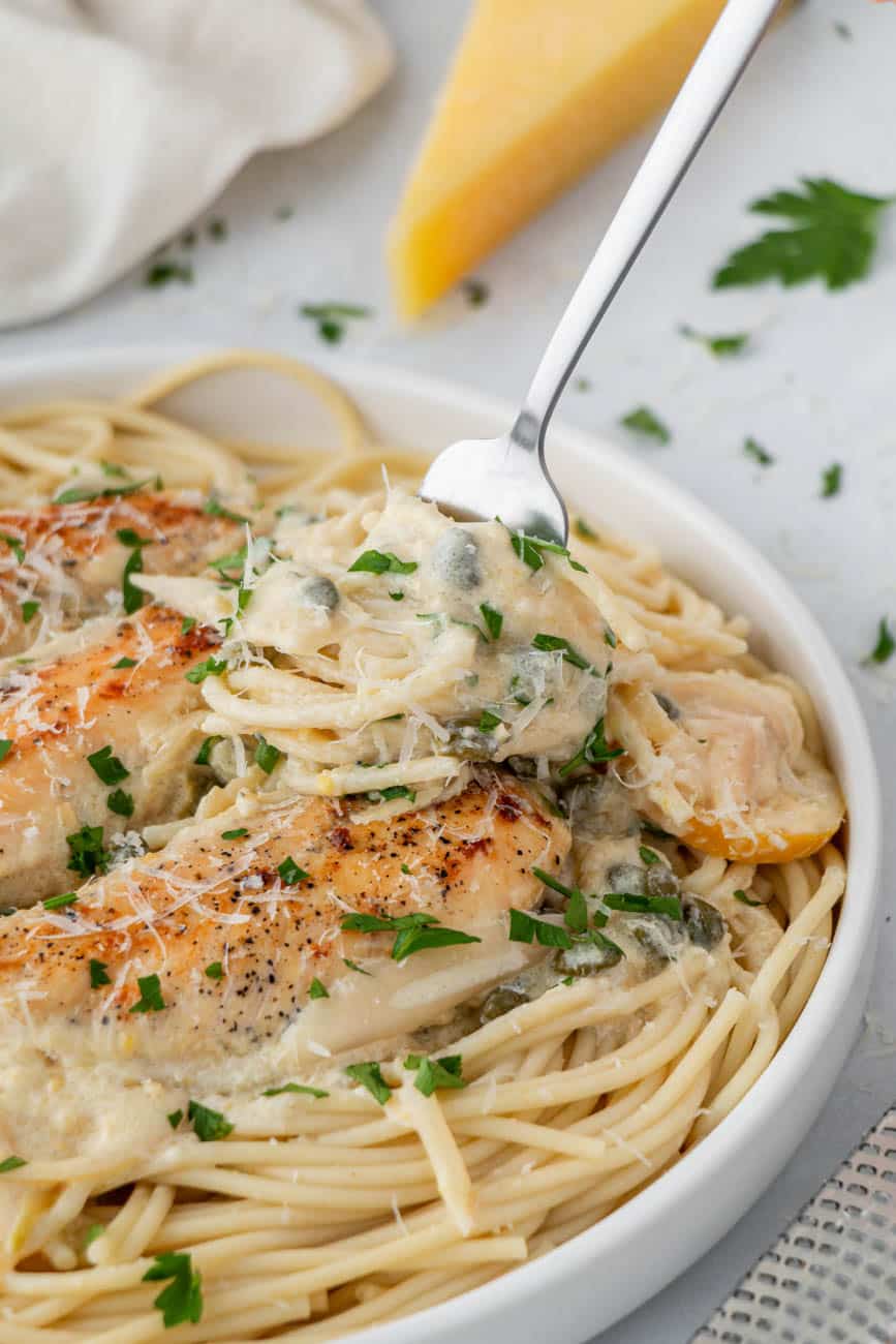 Chicken piccata with pasta in a white bowl with a fork grabbing a bite.