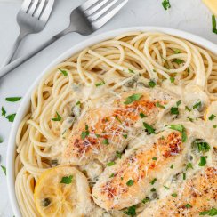 chicken piccata sauce on chicken over a bowl of pasta with two forks on the side