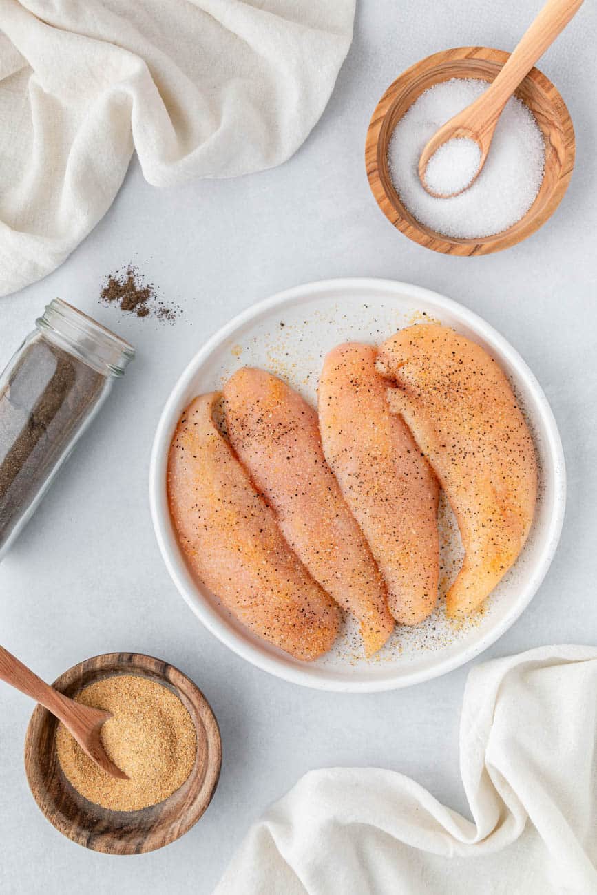 Chicken cutlets on a white plate, pepper, salt, and seasoning.