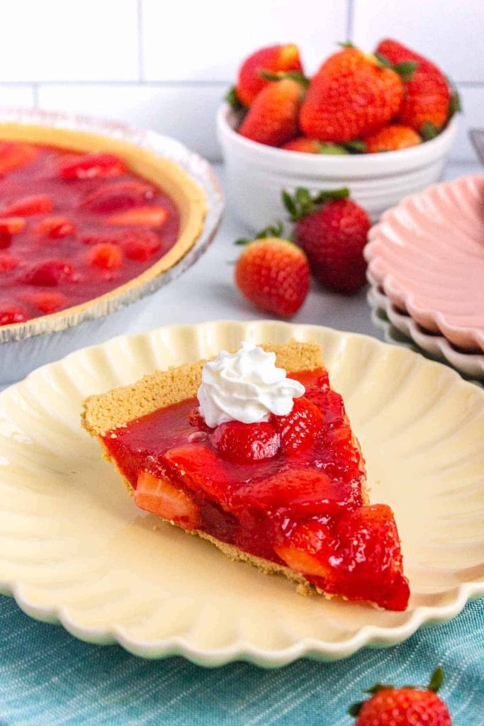 slice of strawberry jello pie topped with whipped cream on a serving plate with more pie in the back