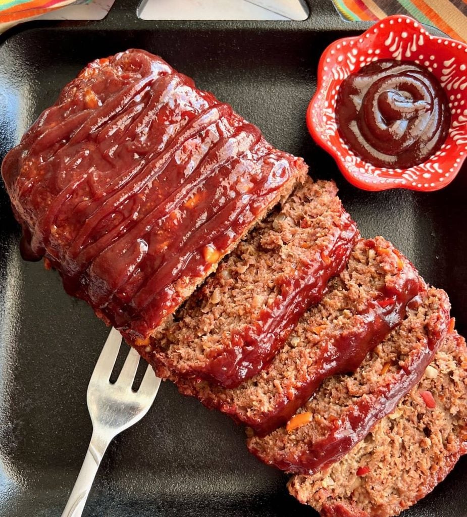 Overhead view of smoked meatloaf with silver fork and sauce in red cup on the side