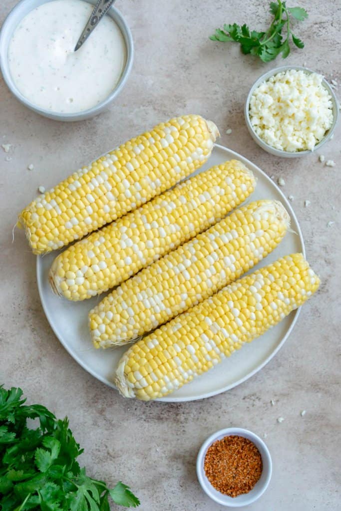 four ears of corn, tajin seasoning, cilantro, cotija cheese, and mexican crema in small ingredient bowls