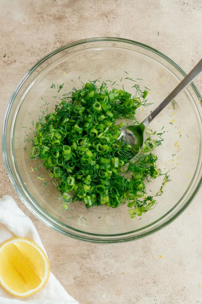 chopped herbs in a glass mixing bowl with a spoon and a sliced lemon on the side.