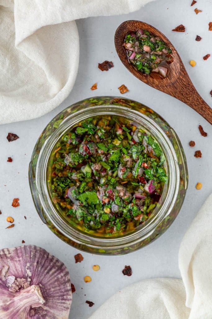 chimichurri sauce in a glass jar with a wooden spoon and garlic on the side