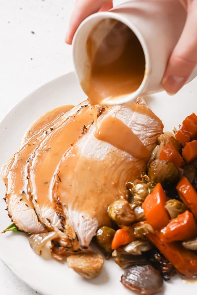 pouring gravy over sliced turkey breast on a white plate with roasted vegetables