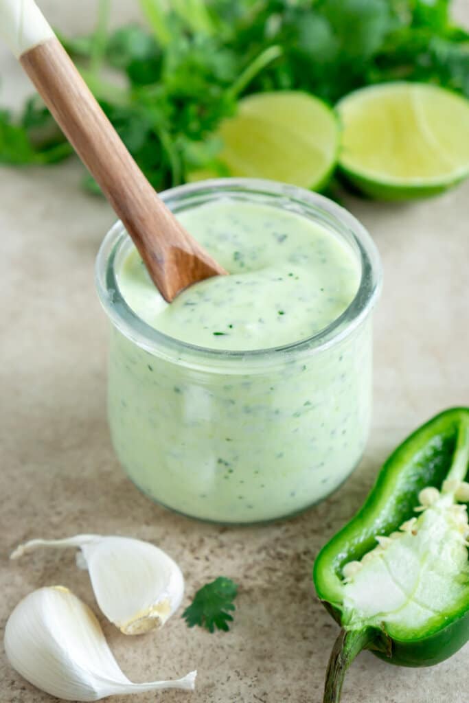a glass jar of jalapeno ranch dressing with a wooden spoon dipped in and garlic, limes, and herbs on the side