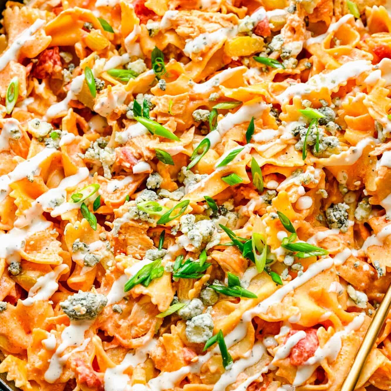 Close up of a skillet full of buffalo chicken pasta, drizzled with blue cheese dressing and garnished with cheese crumbles and green onions.