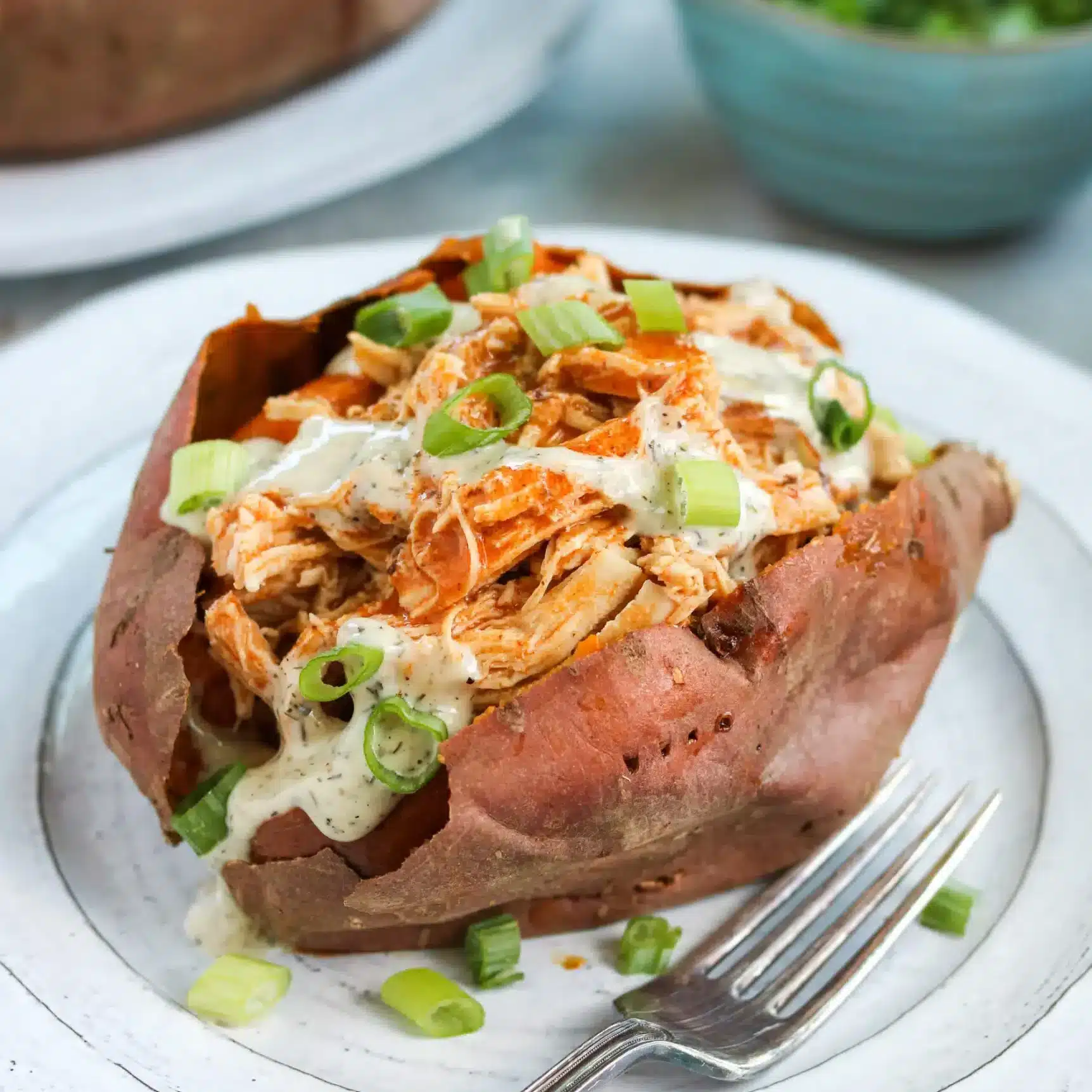 A sweet potato on a plate filled with buffalo chicken and topped with dressing and green onions.