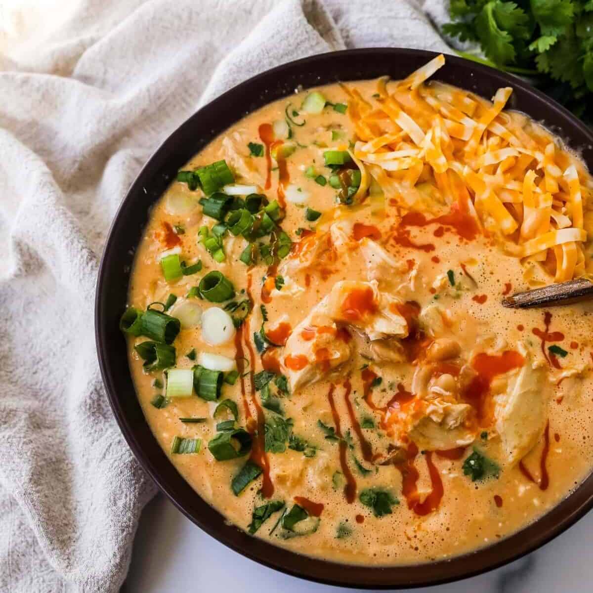 Overhead view of a bowl of buffalo chicken chili with cheese, green onions, and a drizzle of hot sauce.