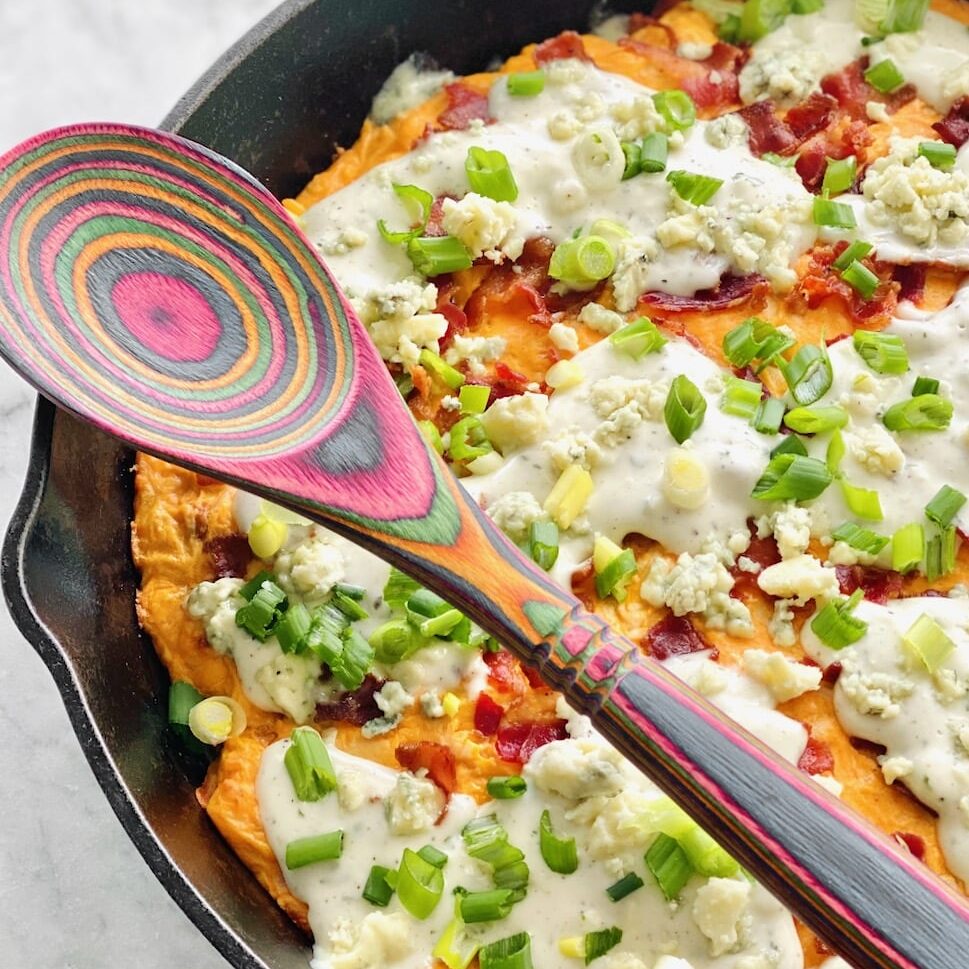 A close up view of a colorful wooden spoon resting across a skillet filled with buffalo chicken tater tot casserole.