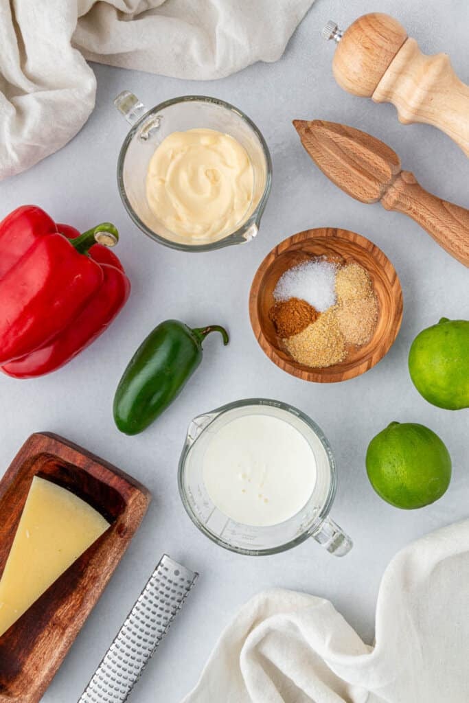 baja sauce ingredients: red pepper, jalapeno, parmesan cheese, spices in a wooden bowl, pepper mill, limes, and a block of cheese