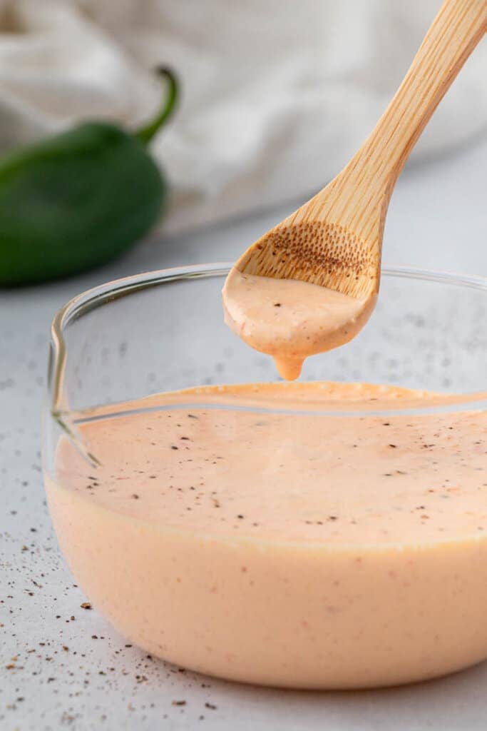 a spoon serving baja sauce from a glass container with a jalapeno and napkin in the background