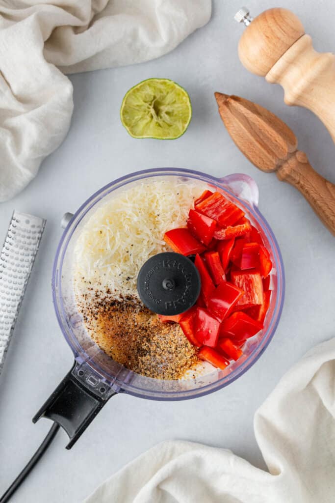 chopped red pepper, parmesan cheese, and spices in a food processor with a squeezed lim and pepper mill to the side