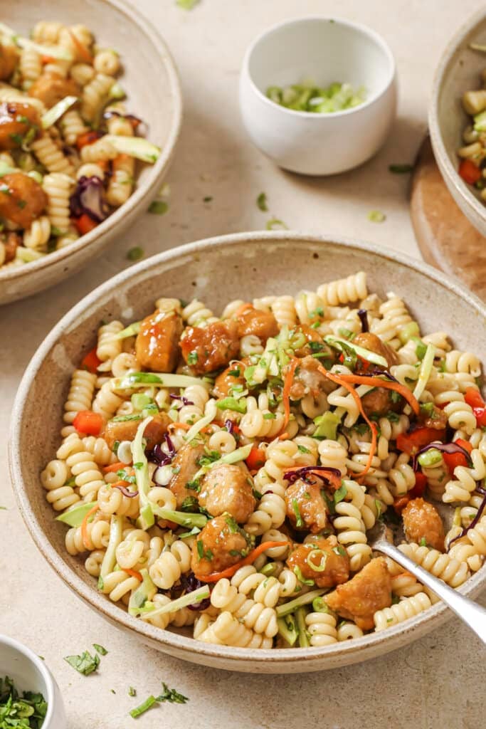 A bowl of orange chicken pasta salad with more servings in the background.