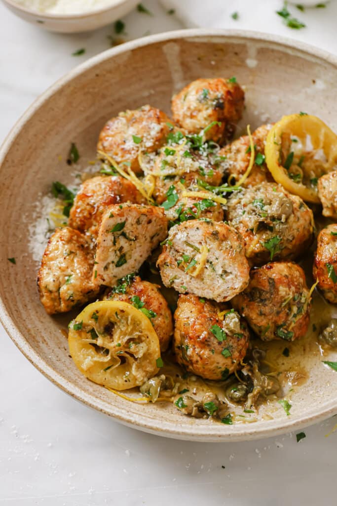 chicken piccata meatballs in an off white bowl, one meatball sliced in half