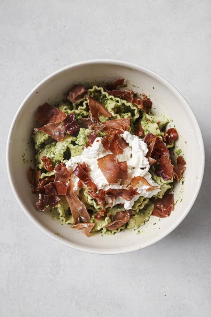 Adding prosciutto and burrata to a bowl of pesto pasta.