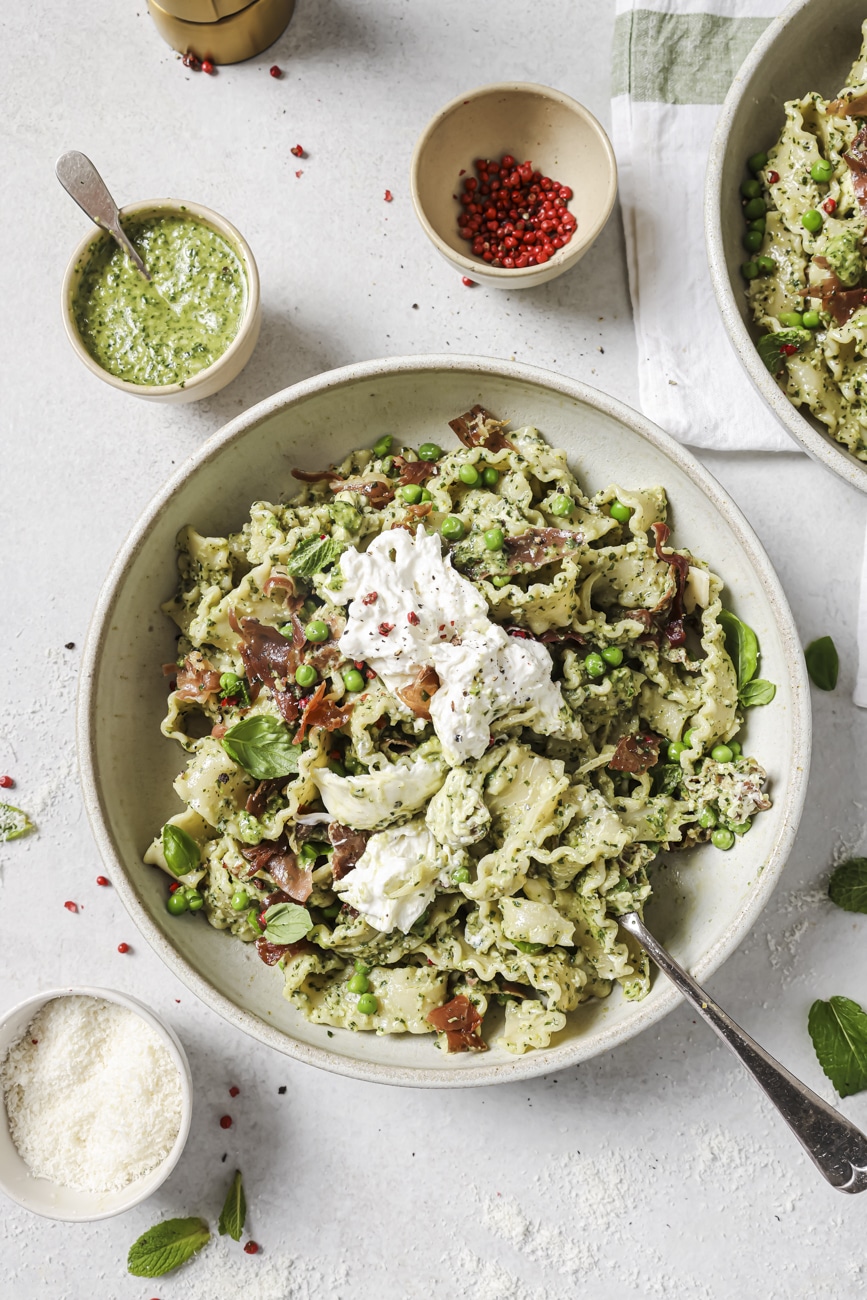 Overhead shot of pesto prosciutto pasta with toppings around the bowl in smaller bowls.