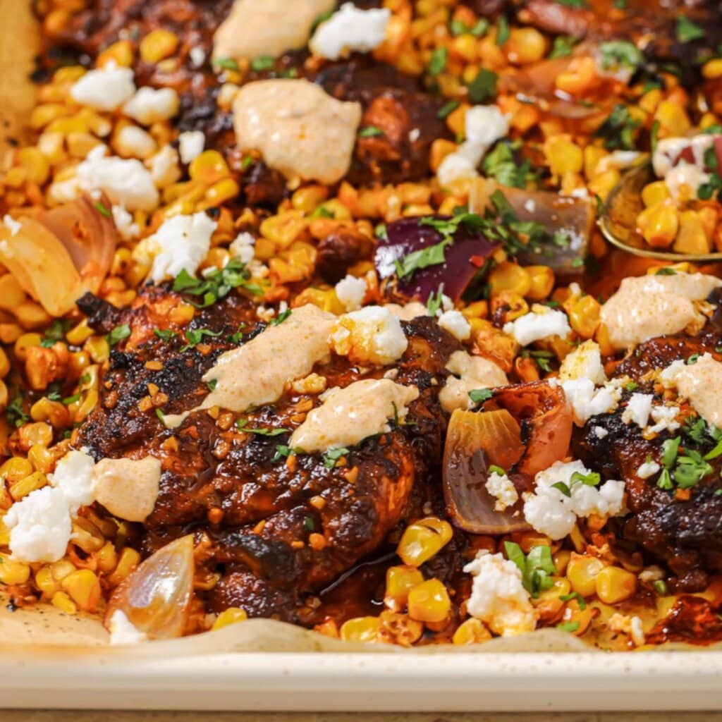 close up photo of sheet pan chicken thighs with corn and sauce drizzled on top.