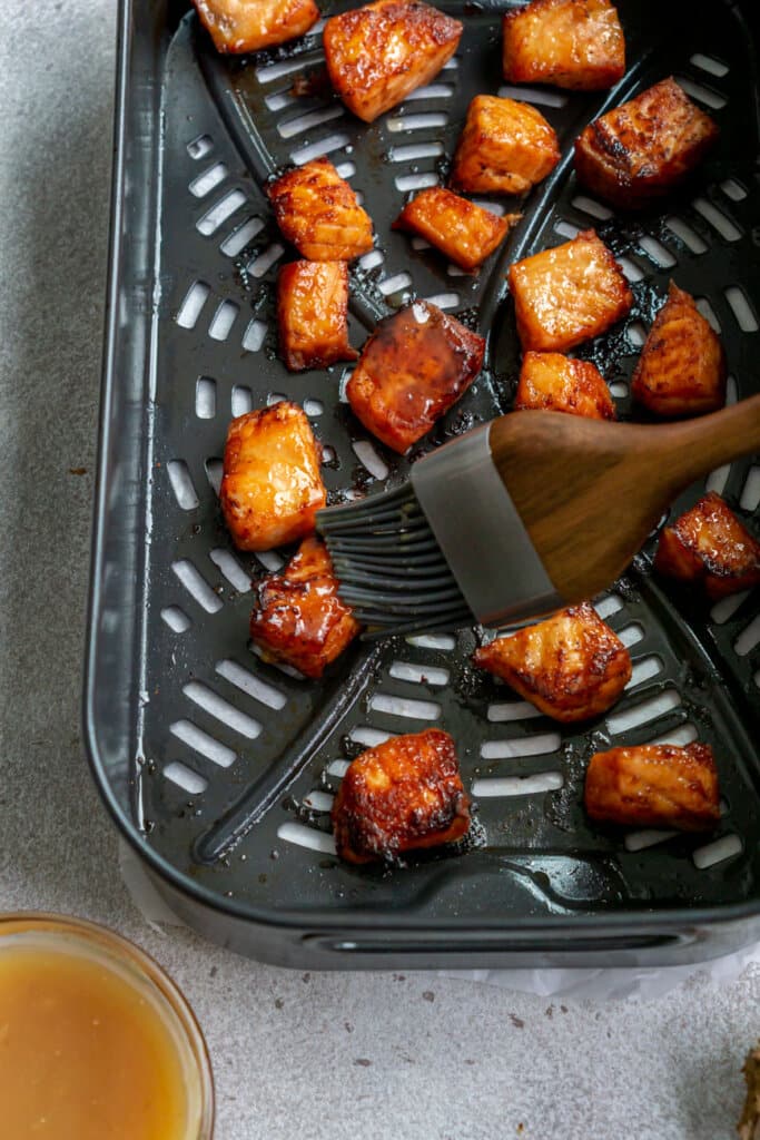 A silicone pastry brush brushes miso glaze onto salmon bites inside an air fryer basket.