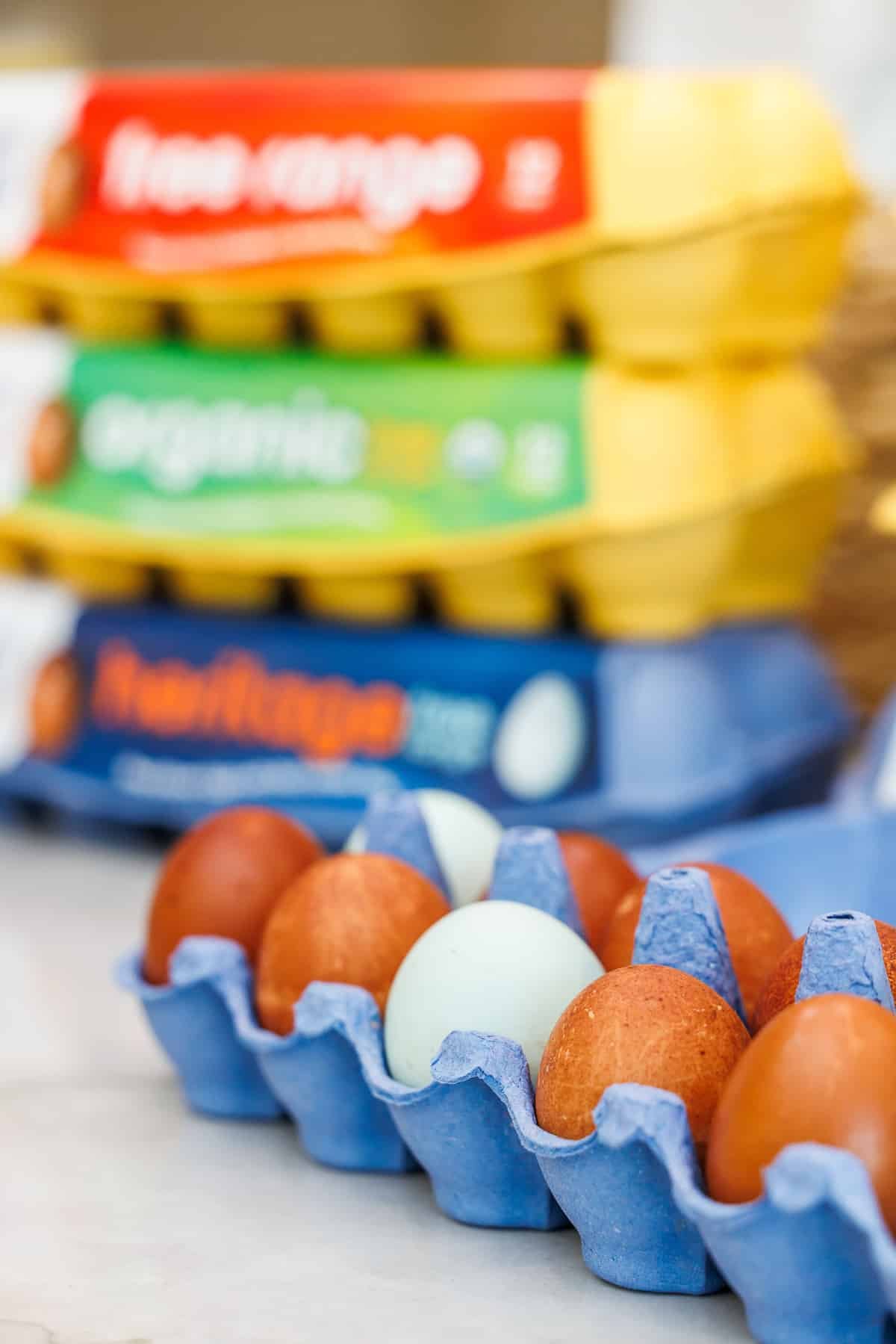 a blue carton of brown eggs, with one blue egg in the middle and a stack of egg cartons in the background.