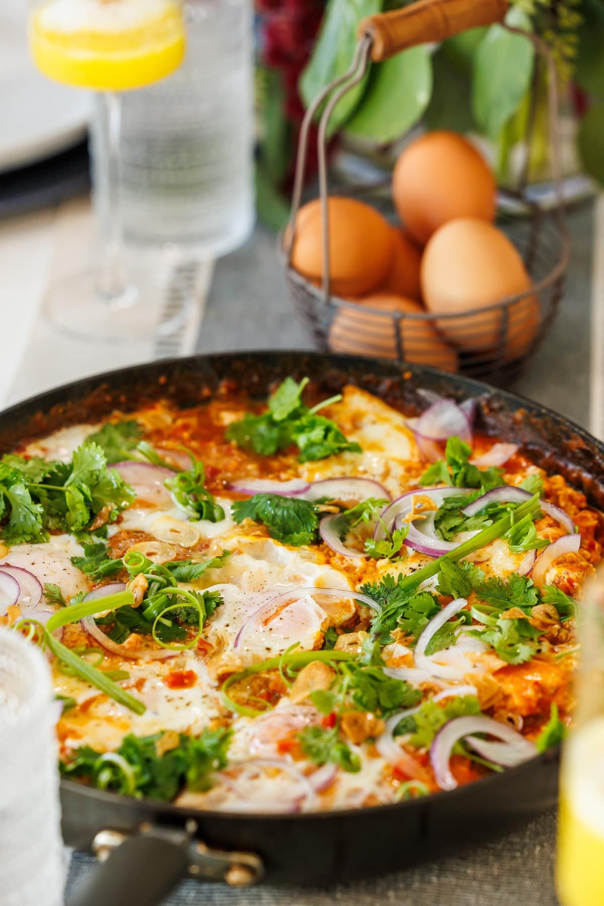 shakshuka in a black cast iron pan with a basket of eggs and a mimosa in the background.