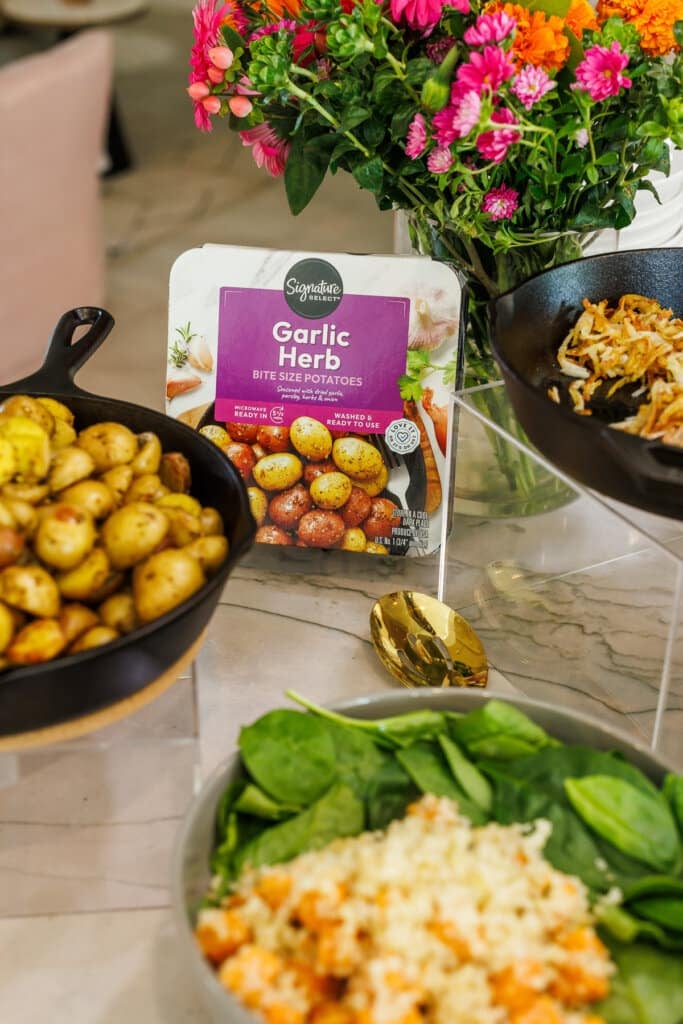 potatoes in a cast iron pan and the package behind them, flowers in the background, and a bowl of spinach and sweet potatoes nearby.