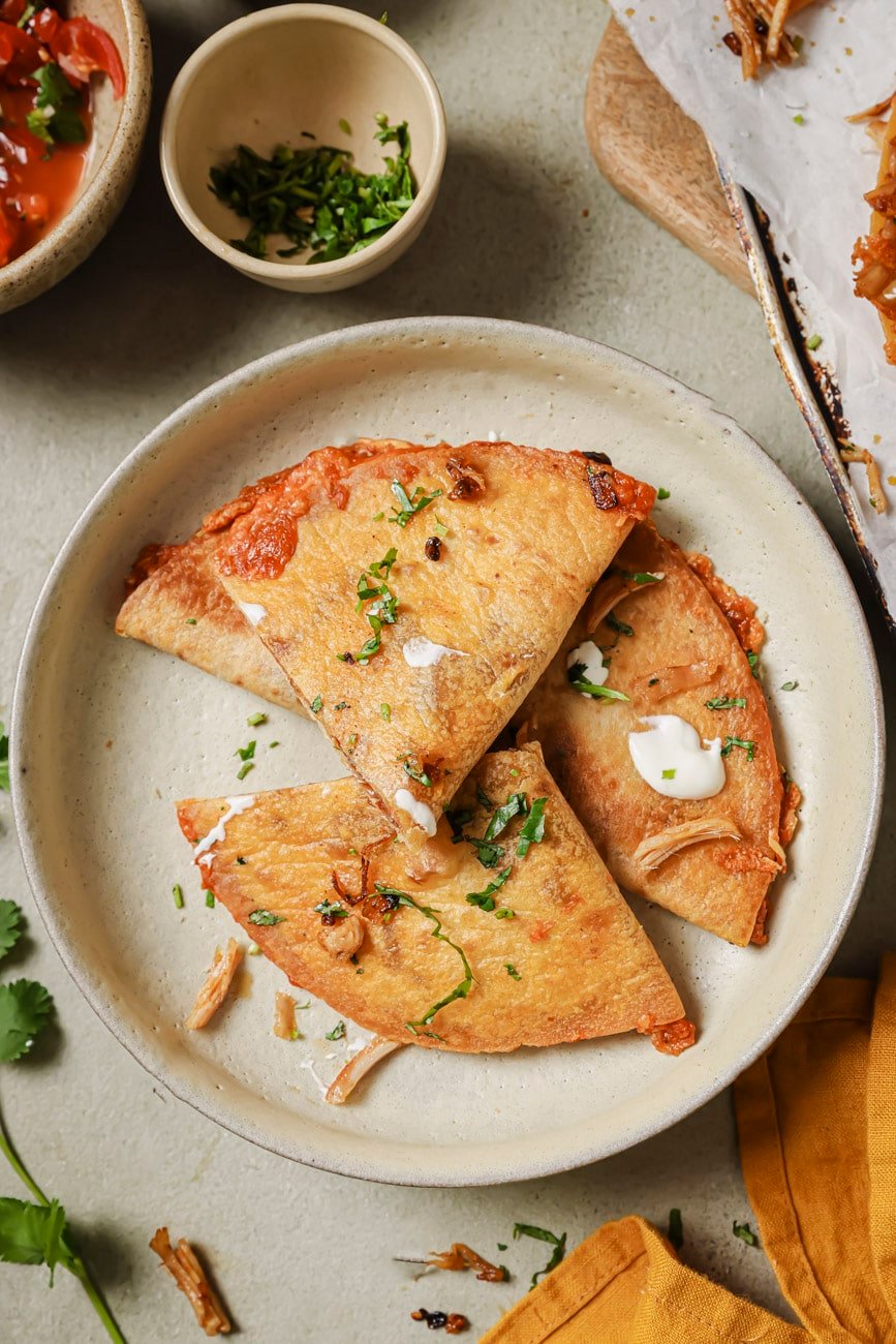 Four turkey quesadillas on a plate with with chopped cilantro on top and a few dollops of sour cream. 