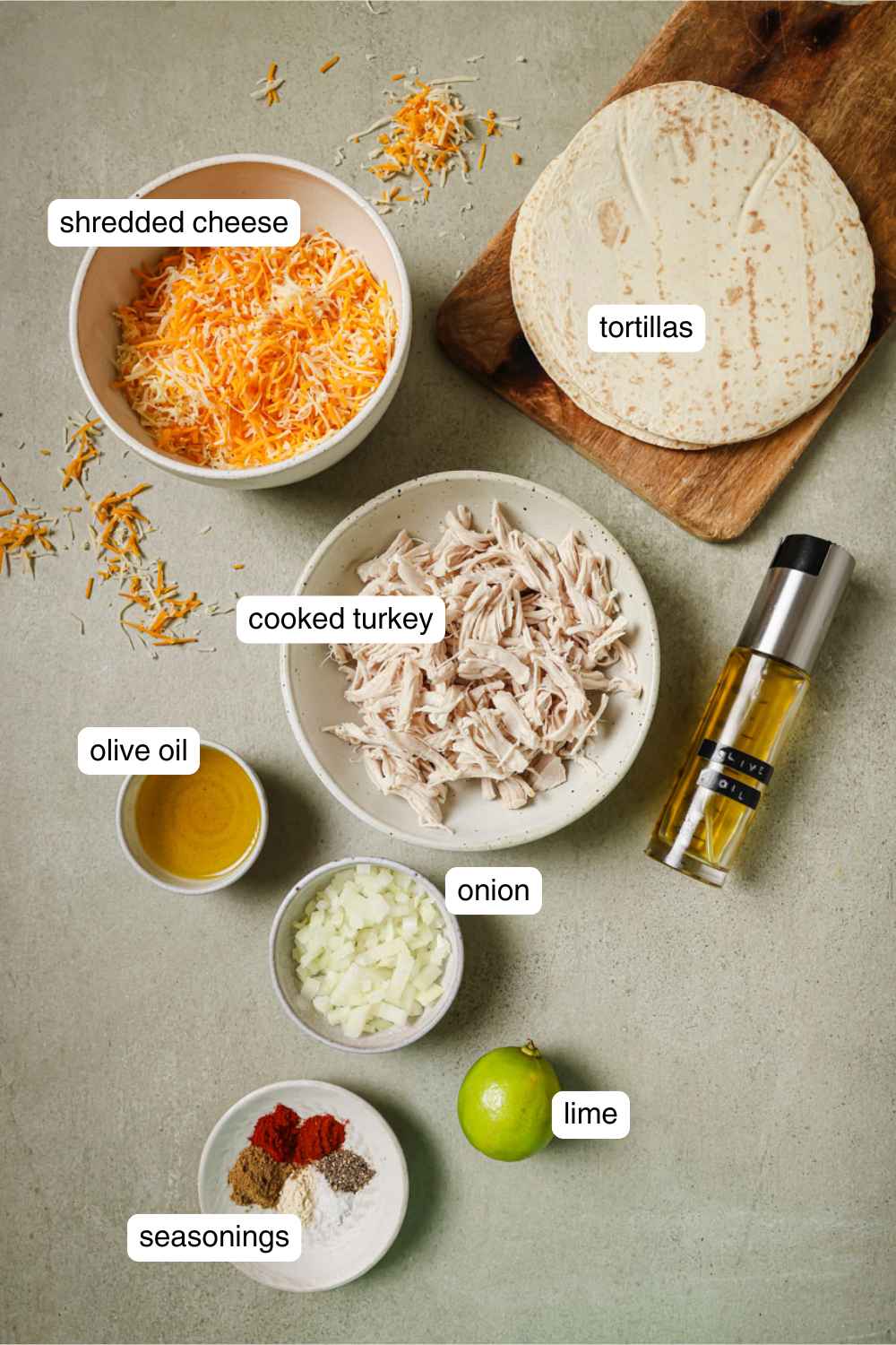 Shredded cheese in a bowl, tortillas on a wood board, cooked turkey in a bowl, olive oil in a small bowl, a lime, seasonings in a small white bowl, and diced onions in a small bowl. 