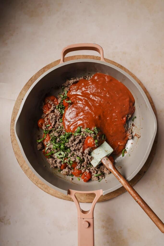 Combining ground beef, sauce, and basil in a pot.