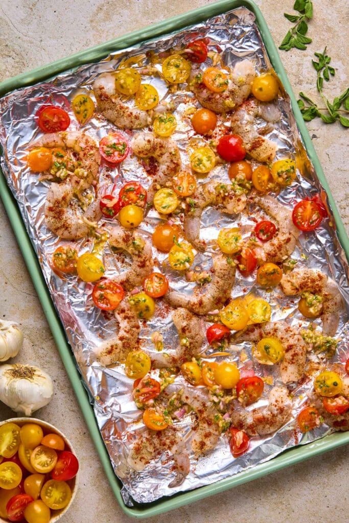 Adding tomatoes to uncooked shrimp on a sheet pan.