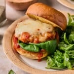 A chicken parmesan burger on a toasted bun with fresh basil on a plate next to fresh greens.