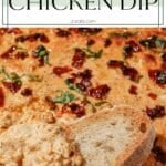 A dish of baked chicken dip topped with sun-dried tomatoes and fresh basil, with a slice of bread dipped into it.