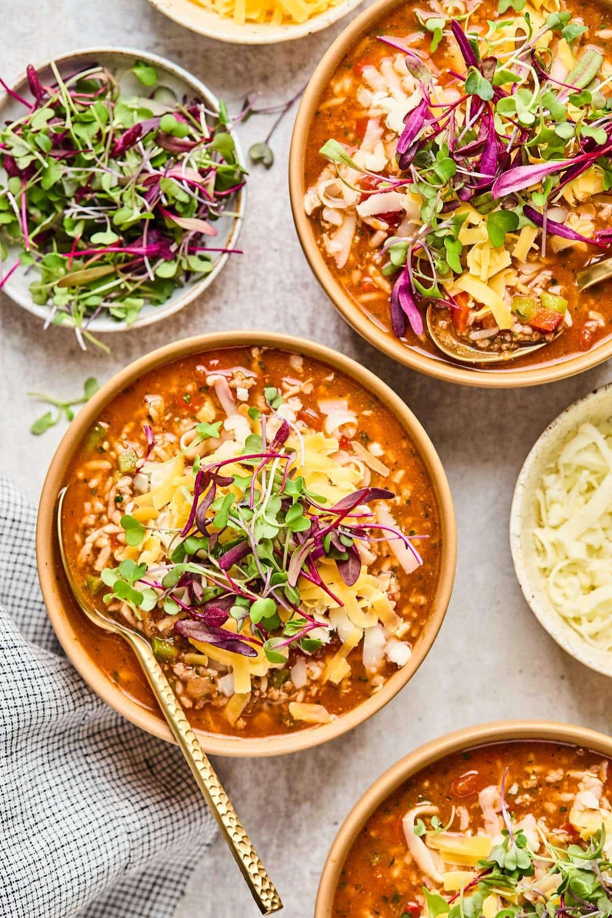 Multiple bowls of stuffed pepper soup atop a counter next to cheese and fresh herbs.