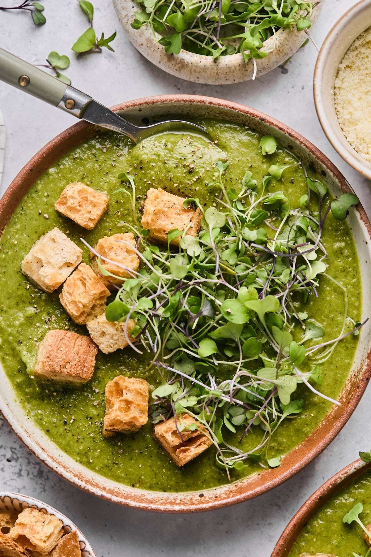 Photo of green soup with fresh greens and croutons on top in a brown bowl with a spoon in the bowl.