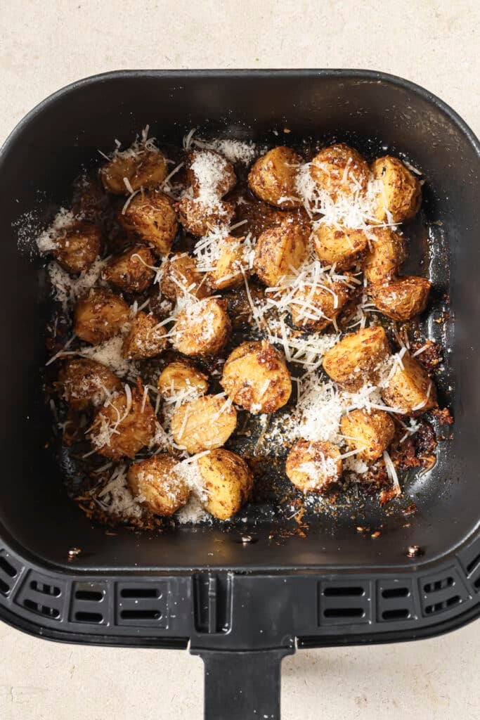 Halved potatoes are in a black air fryer basket. They are topped with grated parmesan cheese.