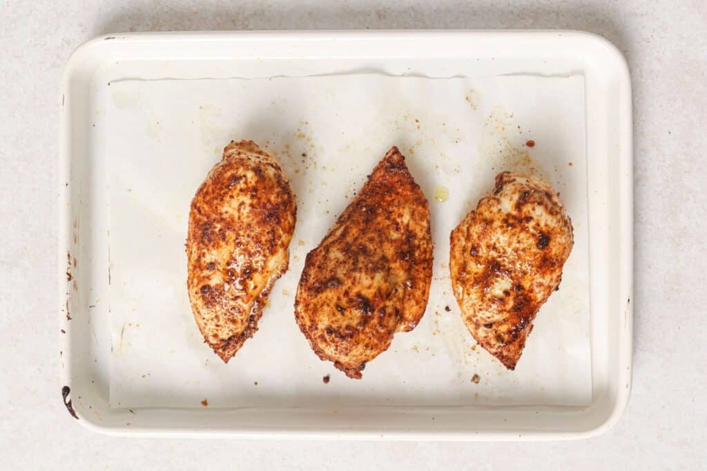 Cooked seasoned chicken breasts on a parchment lined tray.