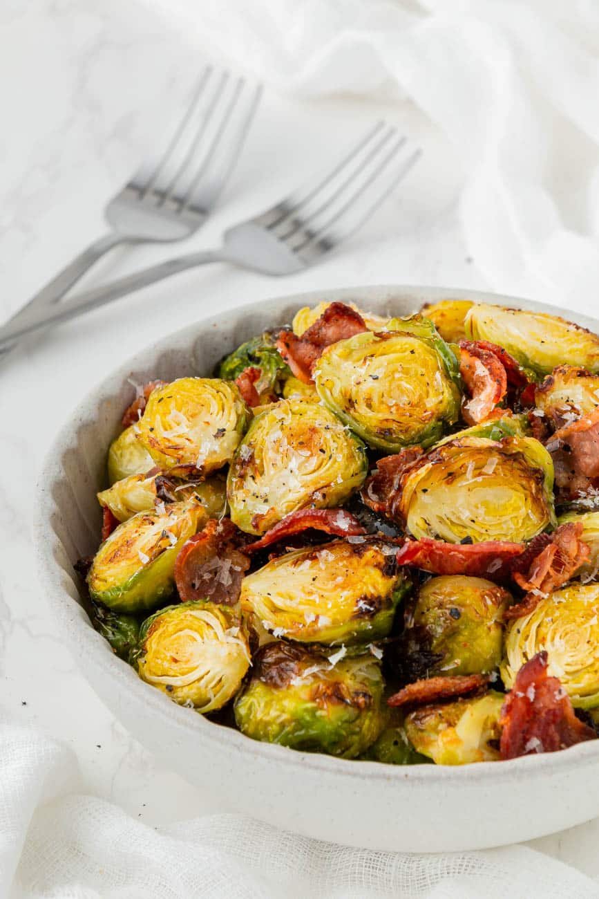 Cooked brussels sprouts and bacon in a white bowl with two forks behind it.