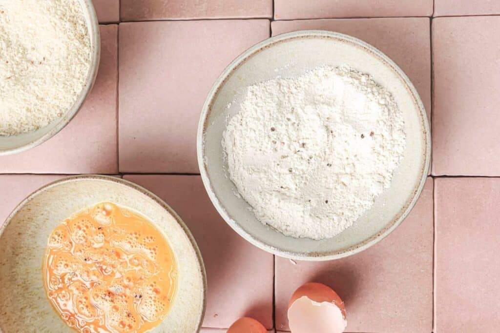 A bowl of flour next to a bowl of beaten egg and a bowl of bread crumbs.