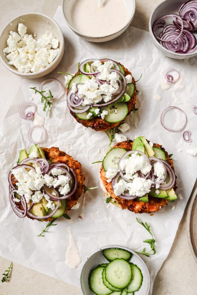 Three open faced salmon burgers with cucumbers, red onion and feta cheese in small bowls on the side.