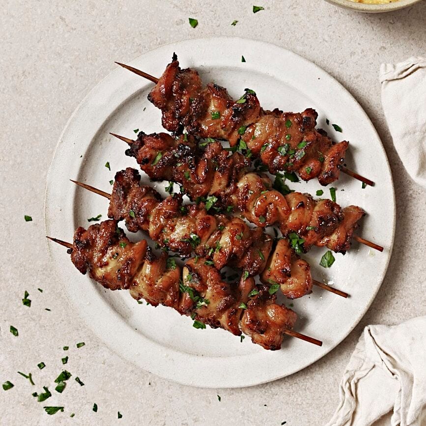 Cooked chicken on four skewers, garnished with parsley and laying on a white plate