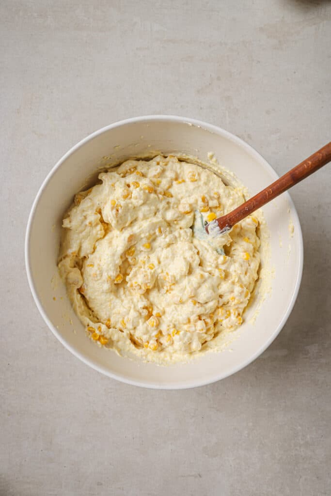 Mixing corn with cornbread and sour cream in a white bowl.