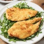 Two baked chicken cutlets on a plate with arugula.