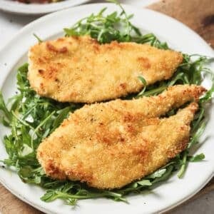 Two baked chicken cutlets on a plate with arugula.