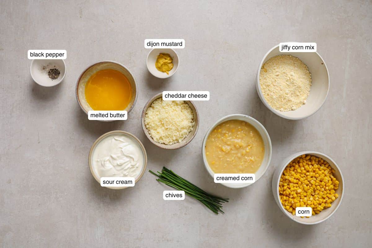Ingredients for baked corn casserole arranged on a light surface, including Jiffy corn mix, creamed corn, corn kernels, cheddar cheese, sour cream, melted butter, Dijon mustard, black pepper, and fresh chives.