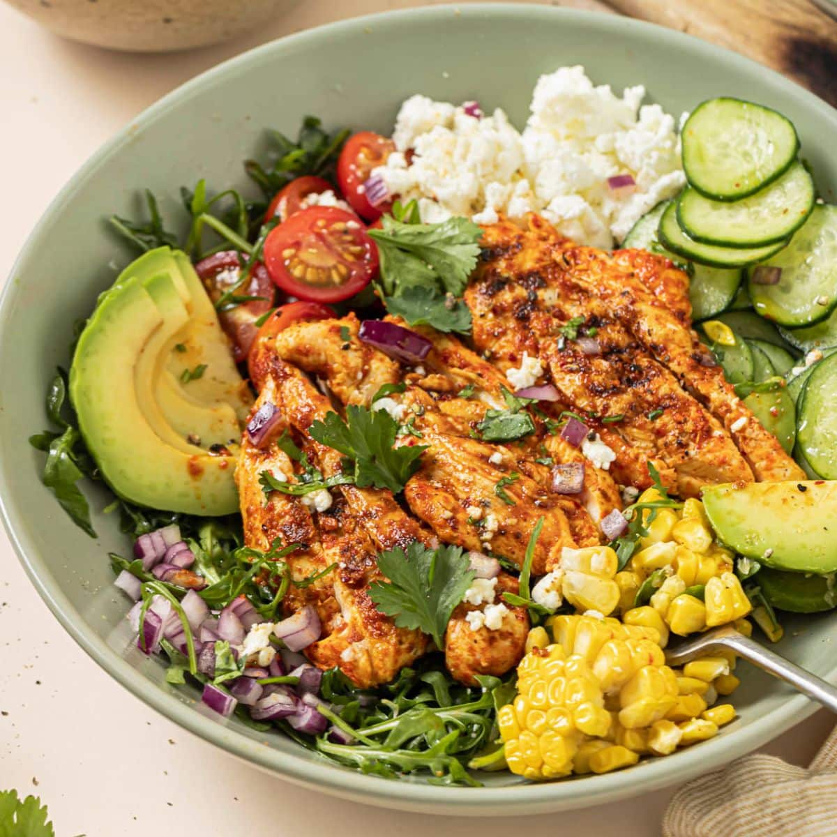 Grilled tajin chicken in a green bowl with corn, cucumbers, feta cheese, and avocado.